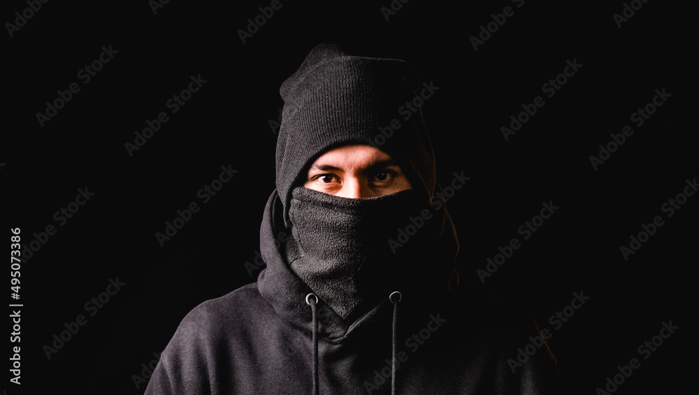 Thief in black balaclava and black cap looking at camera while standing against dark background. Thief in black balaclava and black cap looking at camera while standing against dark background.
