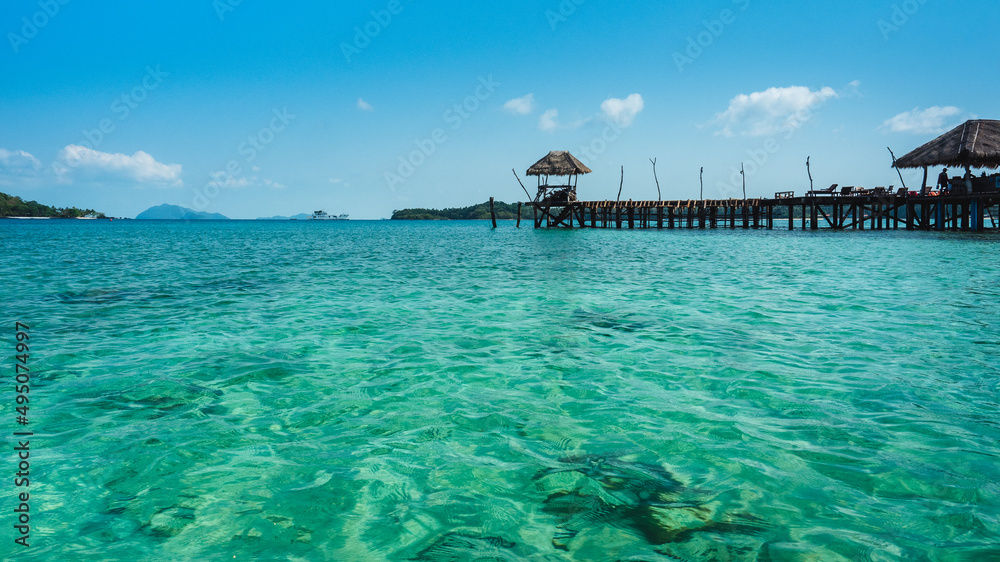 Koh Mak Island, Trat, Thailand. Scenic view of peaceful sea, crystal clear turquoise water with coral reef transparent and long wooden pier bridge landmark against blue sky in summer.
