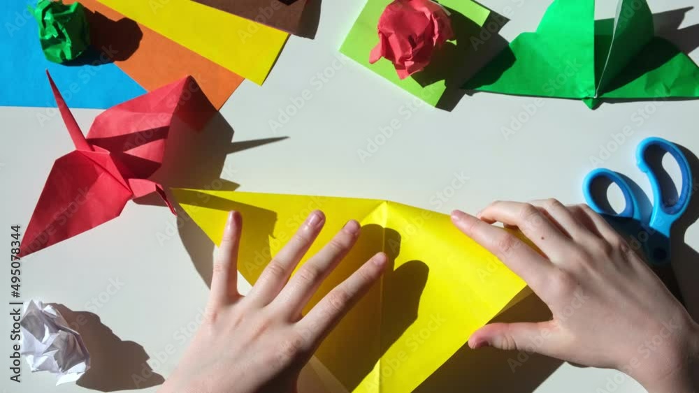 Children's hands do origami from colored paper on white background. Origami paper. DIY. Various school supplies. Concept of children's creativity, back to school.	
