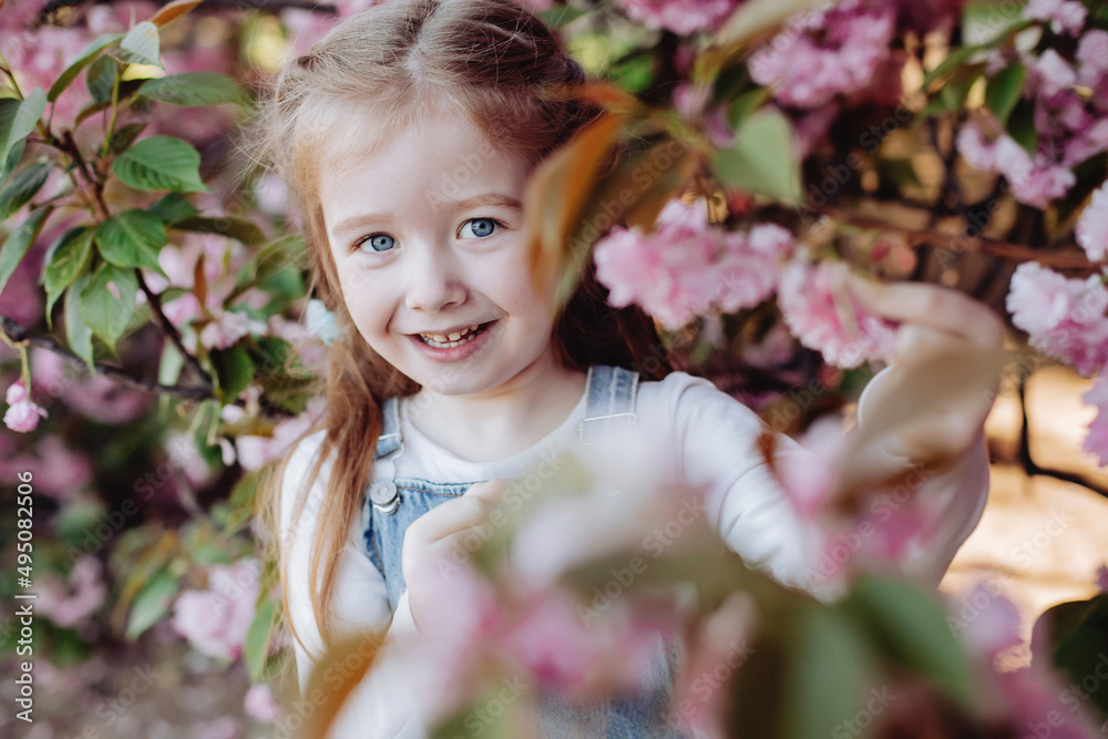 Fototapeta premium Close up of beautiful red-haired girl sincerely smiling in the park near sakura.