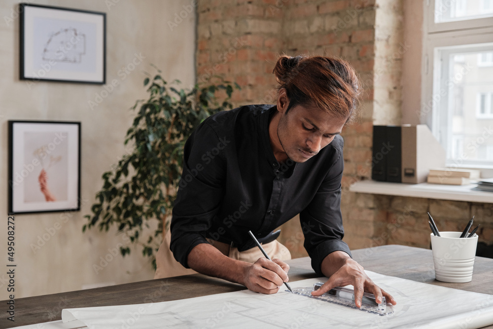 Serious young black architect with ponytail leaning on wooden table and drawing sketch using ruler in loft office Serious young black architect with ponytail leaning on wooden table and drawing sketch using ruler in loft office