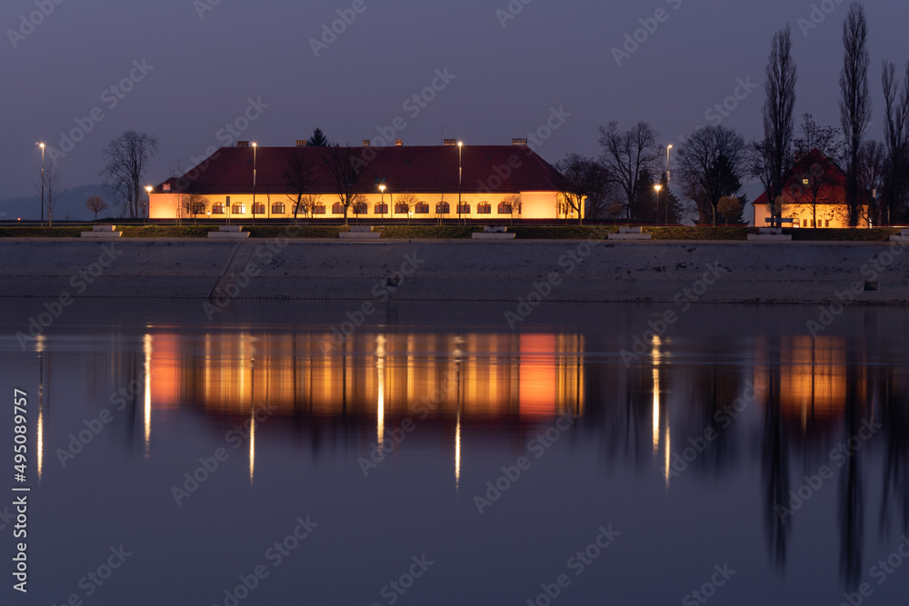 Fototapeta premium The illuminated building of the City Assembly in Slavonski Brod is reflected in the water of the Sava River at night in Croatia