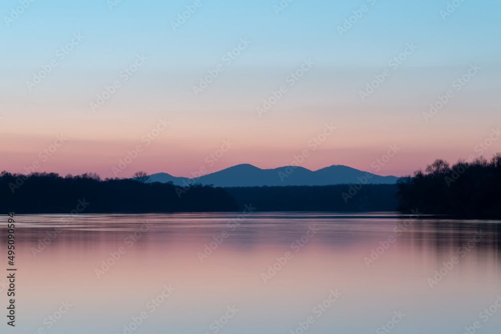 Landscape with river, riparian forest silhouette and distant mountain under clear blue sky with pale red glow at horizon during twilight, pastel colors