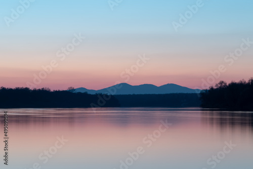 Landscape with river, riparian forest silhouette and distant mountain under clear blue sky with pale red glow at horizon during twilight, pastel colors © slobodan