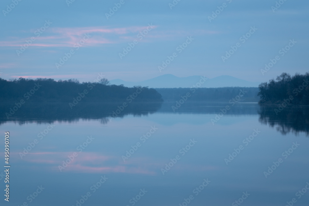Peaceful atmospheric landscape with pastel colors, Sava river at twilight, forested banks lead to distant mountain silhouette in haze, calm nature and water reflection