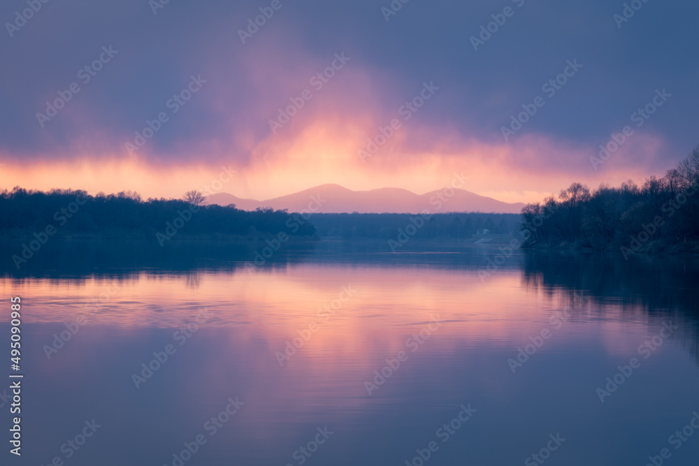 Colorful sunset on Sava river with low clouds and mountain Motajica in bright glowing mist - rural landscape