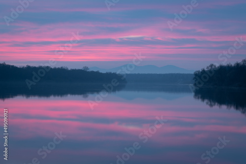 Peaceful atmospheric landscape with pastel colors, Sava river at twilight, forested banks lead to distant mountain silhouette in haze, calm nature and water reflection