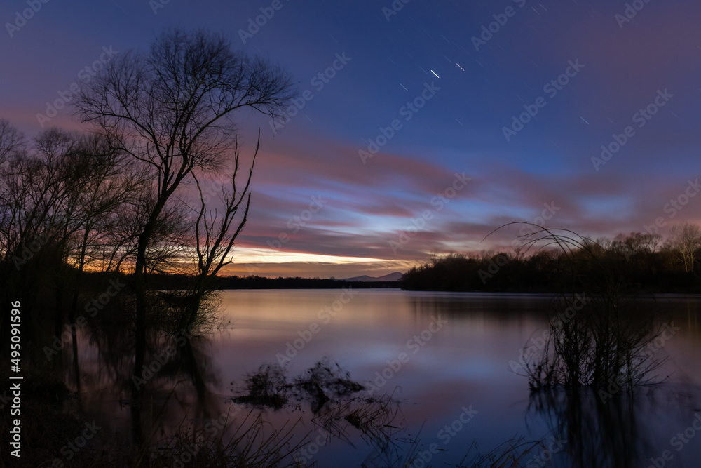 Landscape of Sava river at night in long exposure, star trails in sky and flat water surface, tree silhouette against deep blue sky