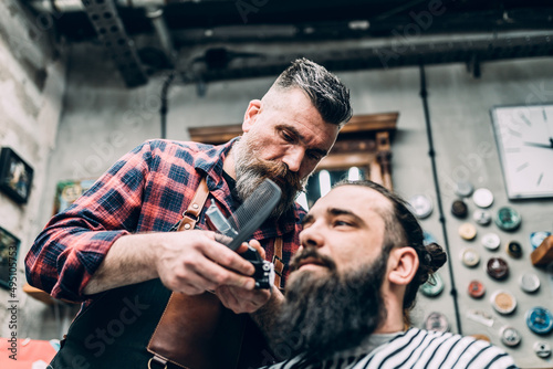 The barber trims the beard of the customer with shaving machine.
