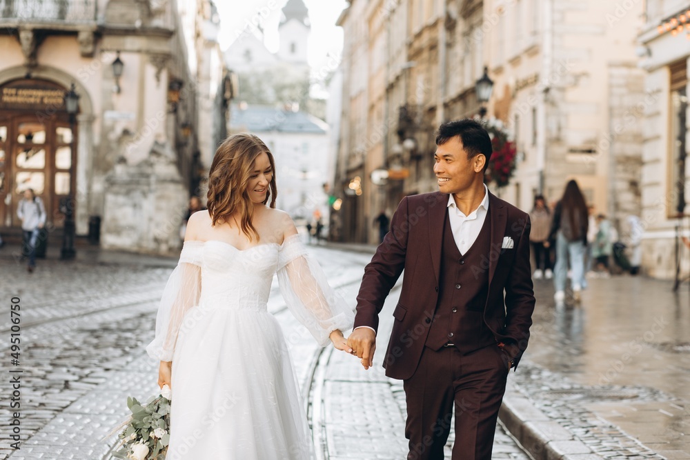 Naklejka premium An international wedding couple, a European bride and an Asian groom walk around the city together.