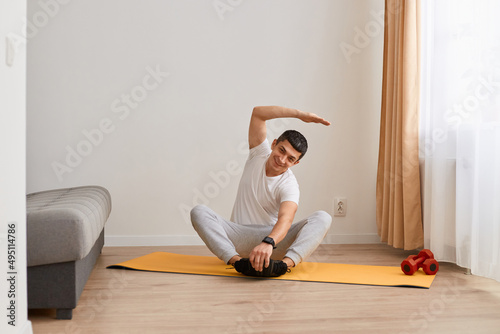 Brunette man wearing sportswear doing sport exercises at home, sitting in lotus position on mat practicing yoga, bending to side with raised hand, doing stretching workouts.