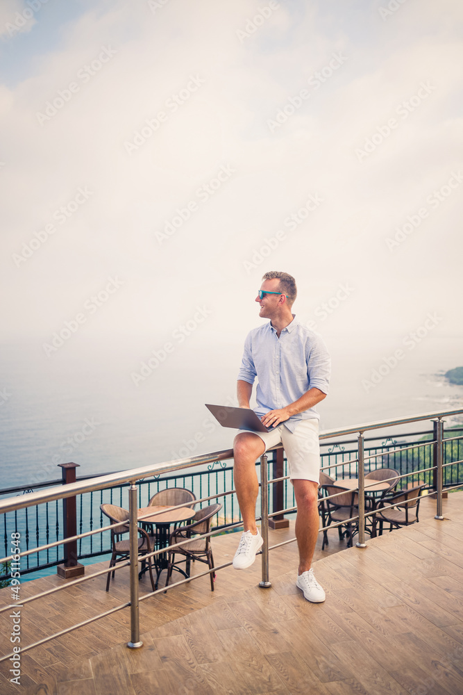 Handsome successful young male businessman working with laptop looks at the Mediterranean Sea. He is wearing a shirt and white shorts. Remote work on vacation. Vacation concept