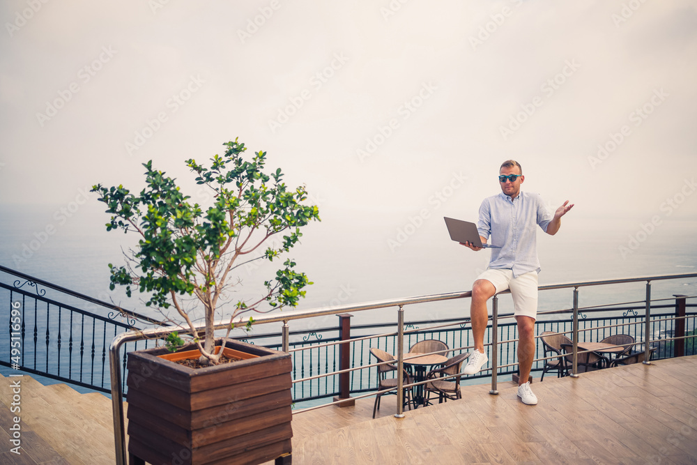 Handsome successful young male businessman working with laptop looks at the Mediterranean Sea. He is wearing a shirt and white shorts. Remote work on vacation. Vacation concept Handsome successful young male businessman working with laptop looks at the Mediterranean Sea. He is wearing a shirt and white shorts. Remote work on vacation. Vacation concept