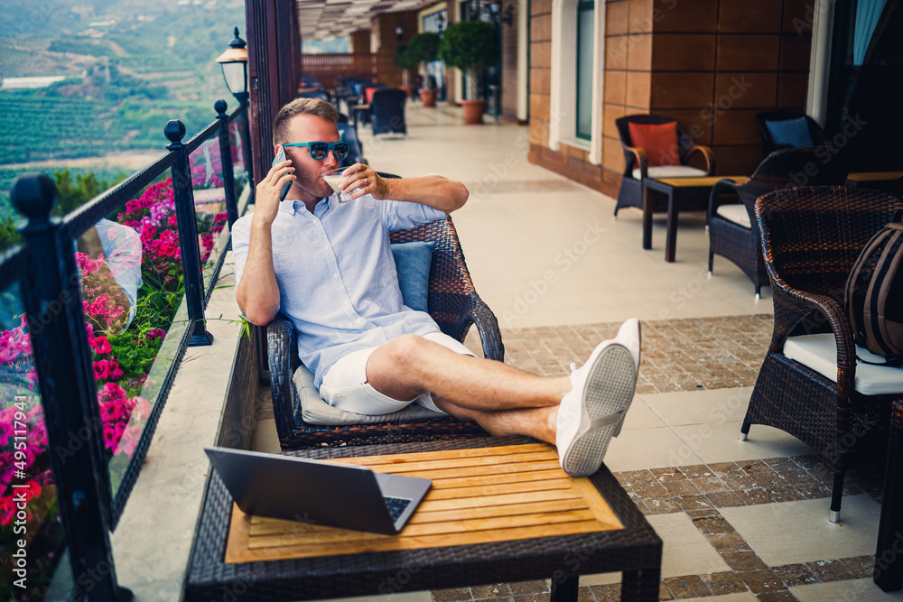 Freelancer concept. Successful young man, businessman working on a laptop computer, sitting on the terrace drinking coffee and talking on the phone.