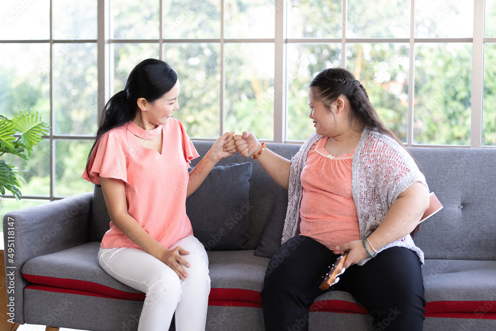 Obraz premium mother giving clashing fists to a girl with down syndrome, singing and playing ukulele or small guitar on sofa