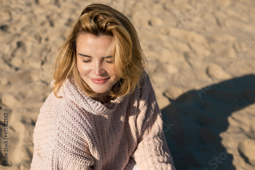 Beautiful girl in sweater sitting on sandy beach at sunny but cold day © leszekglasner