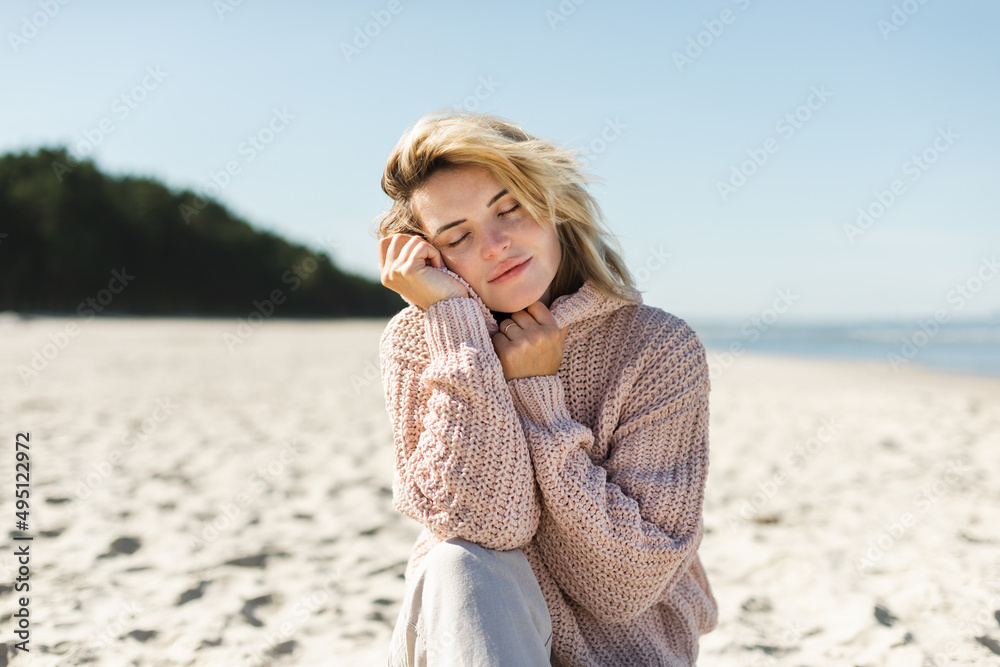 Charming young woman in knitted sweater sitting on sandy beach at sunny day Charming young woman in knitted sweater sitting on sandy beach at sunny day