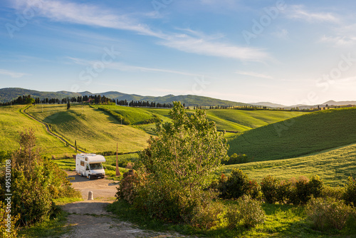 Camper van in unique green landscape, vanlife in Tuscany, Italy. Scenic dramatic sky and sunset light over cultivated hill range and cereal crop fields. Toscana, Italia.