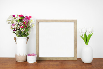 Mock up square wood frame with vases of cut flowers and succulent plant. Wooden shelf against a white wall. Copy space.