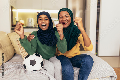 Football match watching concept. Muslim woman and young daughter sit couch cozy celebrate soccer team victory.