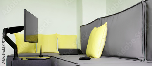 Computer monitor is installed next to gray loft sofa, on small table, using gas-lifted metal swivel bracket. Next to laptop, graphic tablet and mouse. Home workplace for designer or stoker. photo