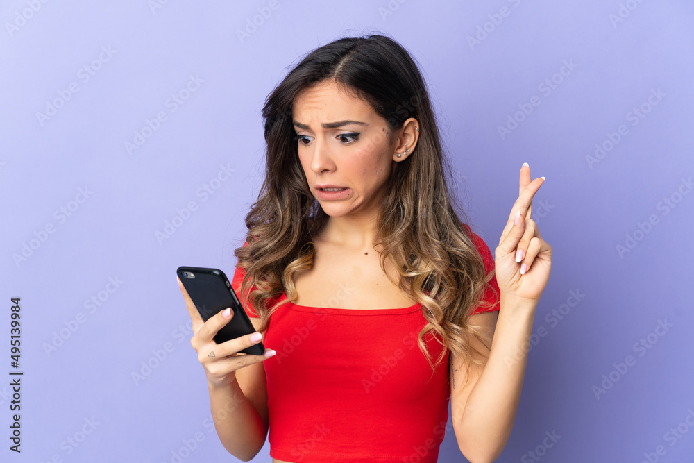 Young caucasian woman isolated on purple background using mobile phone with fingers crossing