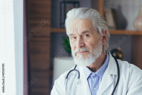 photo close-up portrait of a senior and experienced doctor, gray-haired man looking into the camera, working in a private office, dressed in a medical gown