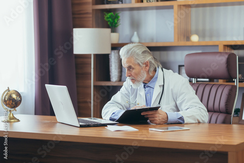 Gray-haired experienced male doctor working on laptop remotely advising patients while sitting in office