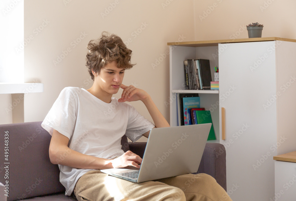 Cute young teenager with laptop on the couch at home. Boy makes homework with a computer. Distance, online education for children Cute young teenager with laptop on the couch at home. Boy makes homework with a computer. Distance, online education for children