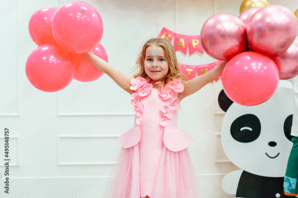 Portrait of little smiling happy girl with long wavy fair hair and painted face wearing pink dress decorated with flowers, holding pink balloons, spreading arms near cardboard panda at birthday party. Portrait of little smiling happy girl with long wavy fair hair and painted face wearing pink dress decorated with flowers, holding pink balloons, spreading arms near cardboard panda at birthday party.