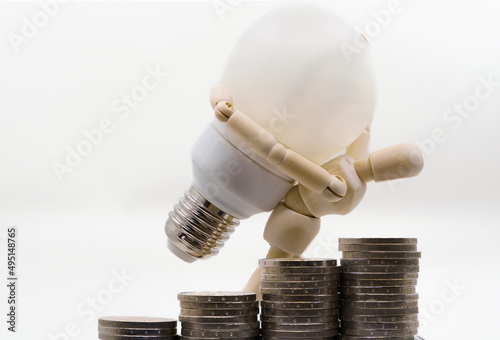 wooden doll holding a light bulb on his shoulders with a rising coin graph in the foreground . Concept of rising electricity prices