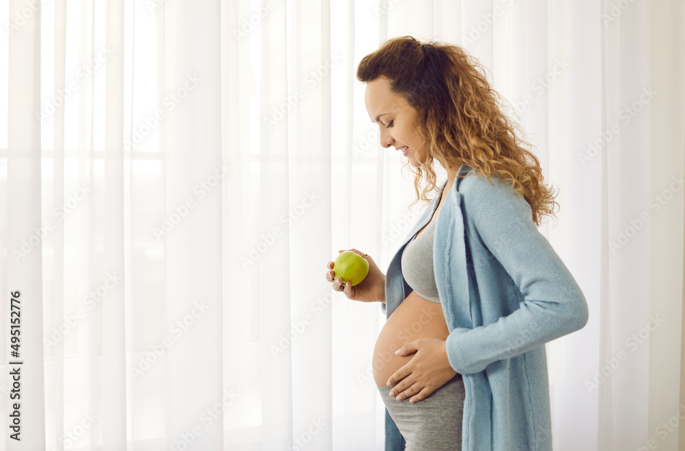 Profile view of a beautiful young pregnant woman with a big belly standing by the window and holding a fresh tasty green apple. Eating healthy food during pregnancy and maintaining good health concept Profile view of a beautiful young pregnant woman with a big belly standing by the window and holding a fresh tasty green apple. Eating healthy food during pregnancy and maintaining good health concept