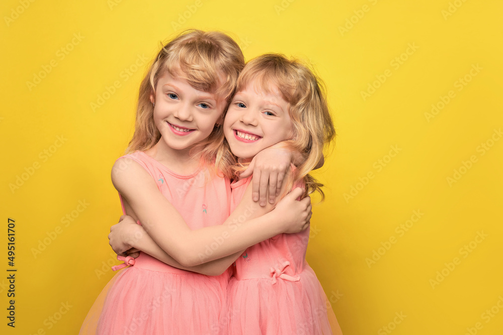 Portrait of twin girls. Two cute attractive cheerful girls of preschool age. Smiling sisters hugging and looking at the camera. Curly blonde 5 years old in a pink dress on a bright yellow background. Portrait of twin girls. Two cute attractive cheerful girls of preschool age. Smiling sisters hugging and looking at the camera. Curly blonde 5 years old in a pink dress on a bright yellow background.