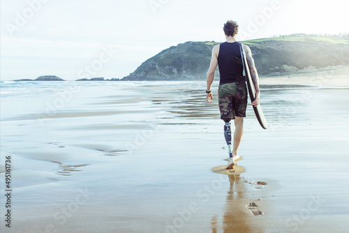 Caucasian surfer man, seen from behind, walking along the beach with an artificial leg.Copy Space