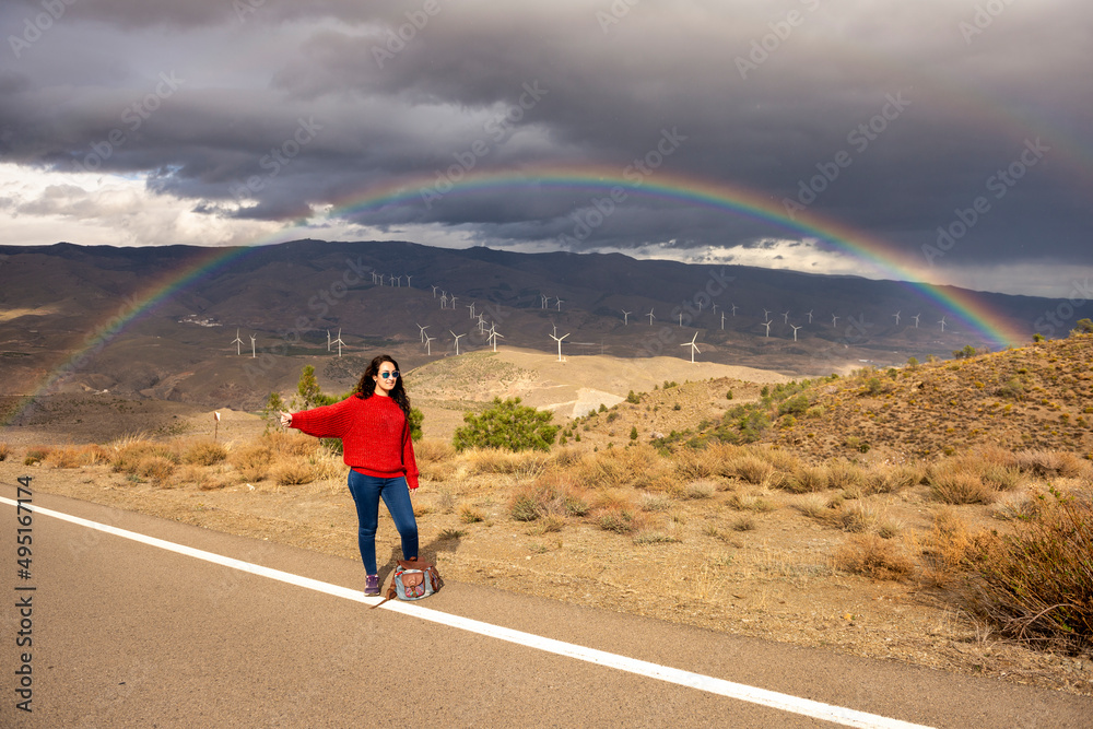 Woman in her 25s hitchhiking on a day with sun, clouds and rainbows in the background.