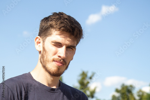Handsome young guy with a beard squints in the sun. Sunshine good weather. Blue sky background