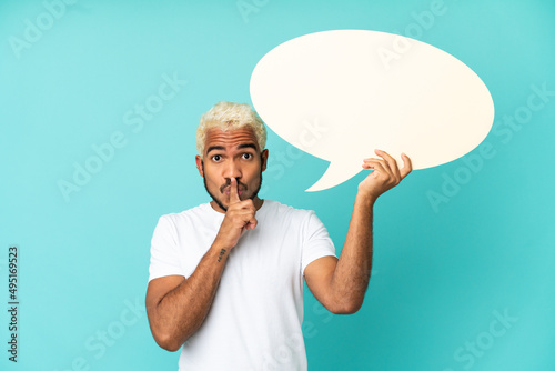 Young Colombian handsome man isolated on blue background holding an empty speech bubble and doing silence gesture