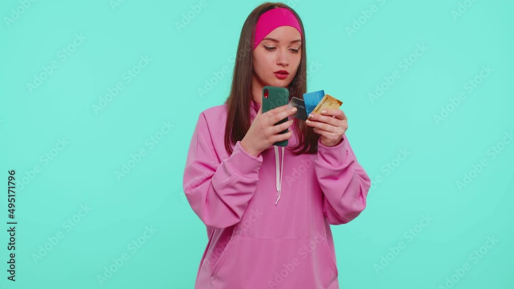 Sincere teen student girl 20s years old in pink hoodie using credit bank cards and smartphone while transferring money, purchases online shopping. Young adult woman isolated on blue wall background Sincere teen student girl 20s years old in pink hoodie using credit bank cards and smartphone while transferring money, purchases online shopping. Young adult woman isolated on blue wall background