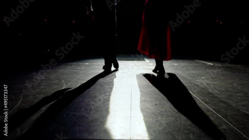 Feet of ballroom dancers pair in shoes and high heels slowly coming out onto proscenium. lit by spotlights in front of dark auditorium.