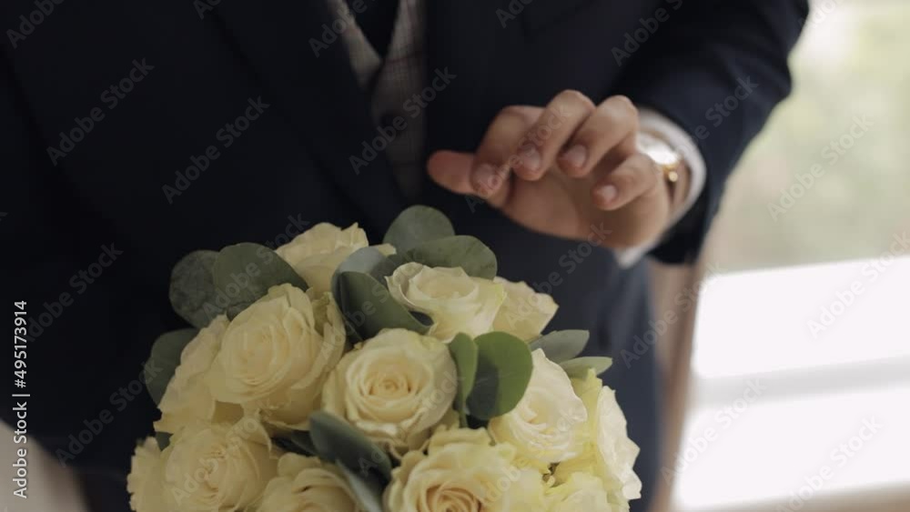 Hands of groom with wedding bouquet at home. White shirt and blue jacket, wrist watch clock. Wedding day. Handsome man preparing to go to bride. Businessman guy. Close-up indoors footage, slow motion Hands of groom with wedding bouquet at home. White shirt and blue jacket, wrist watch clock. Wedding day. Handsome man preparing to go to bride. Businessman guy. Close-up indoors footage, slow motion