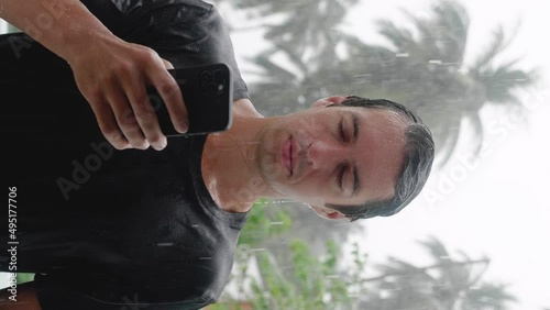 Vetrical shot of wet young man standing in the pouring tropical rain using his smartphone. Slow motion