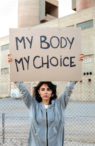 Vertical portrait of young caucasian woman looking at camera holding cardboard sign: My body my choice.