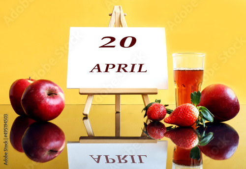 Calendar for April 20: an easel with the inscription 20, april, apples and strawberries next to it, a glass of freshly squeezed juice, yellow background, side view
