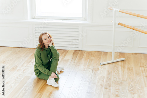 Female dancer has rest after training, poses on floor near ballet barre, has ballet classes, wears green sweatsuit, sportshoes, likes being active