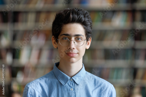 Head shot schoolboy guy posing in campus library on bookshelves background. 17s pupil in eyeglasses look at cam. Excellent student portrait, skill and knowledge, education, generation Z person concept photo