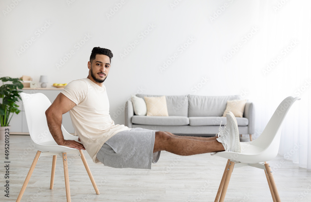 Athletic young Arab man doing strength exercises with two chairs at home