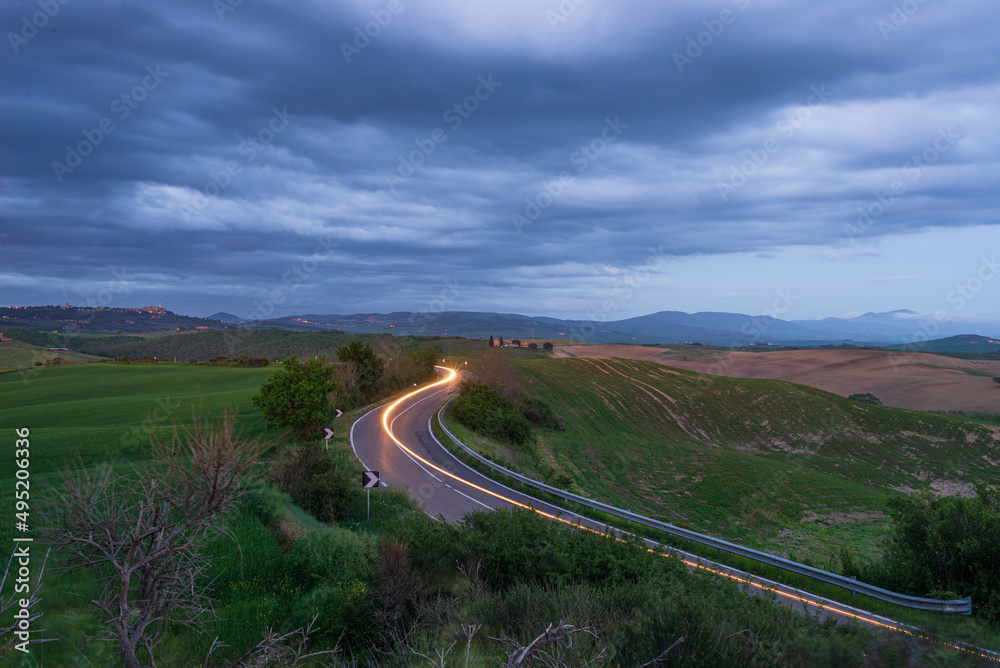Car light trail on winding road in the unique hill landscape of Tuscany, Italy. Dramatic sunset sky at twilight, illuminated villages on hilltop.