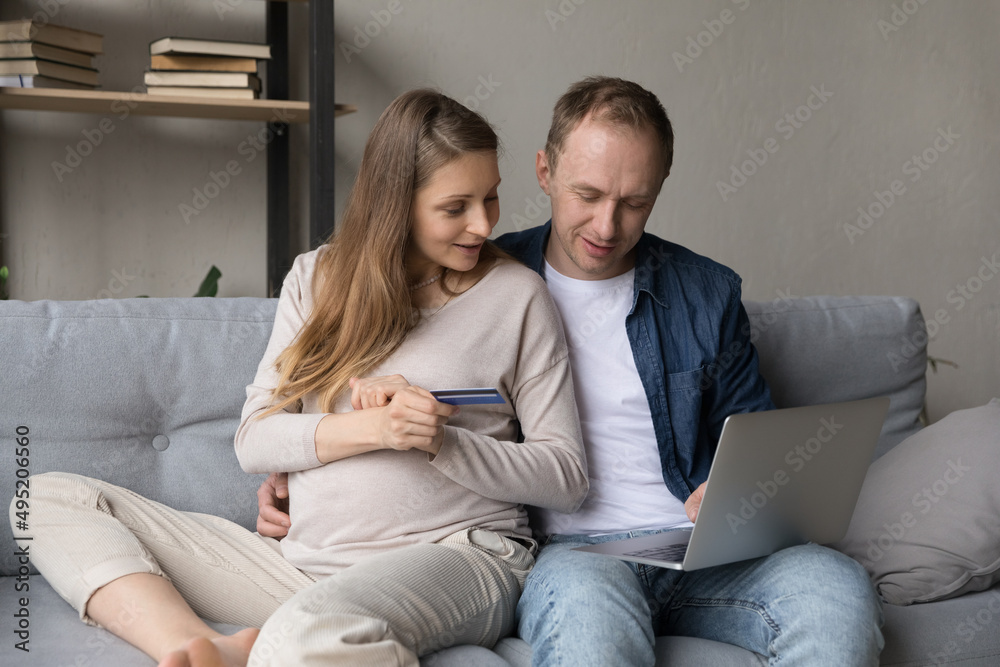 Happy beautiful young pregnant woman holding bank credit card in hands, enjoying online shopping with affectionate husband using computer, choosing goods for future baby, preparing for childbirth.