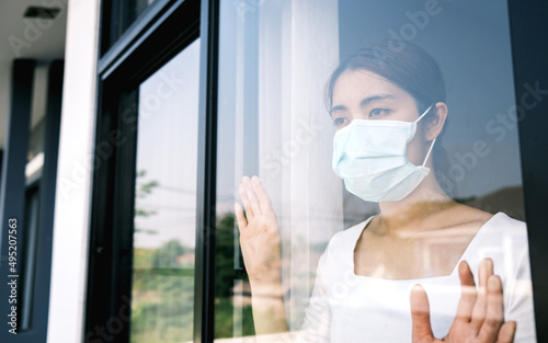 Young woman wearing a medical mask looking through the window. Patient isolated to prevent infection due to epidemic of covid 19, coronavirus. Home isolation concept