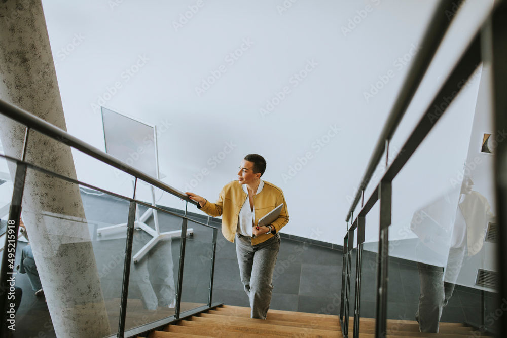 © BGStock72 - Young short hair business woman walking on office stairs and holding laptop